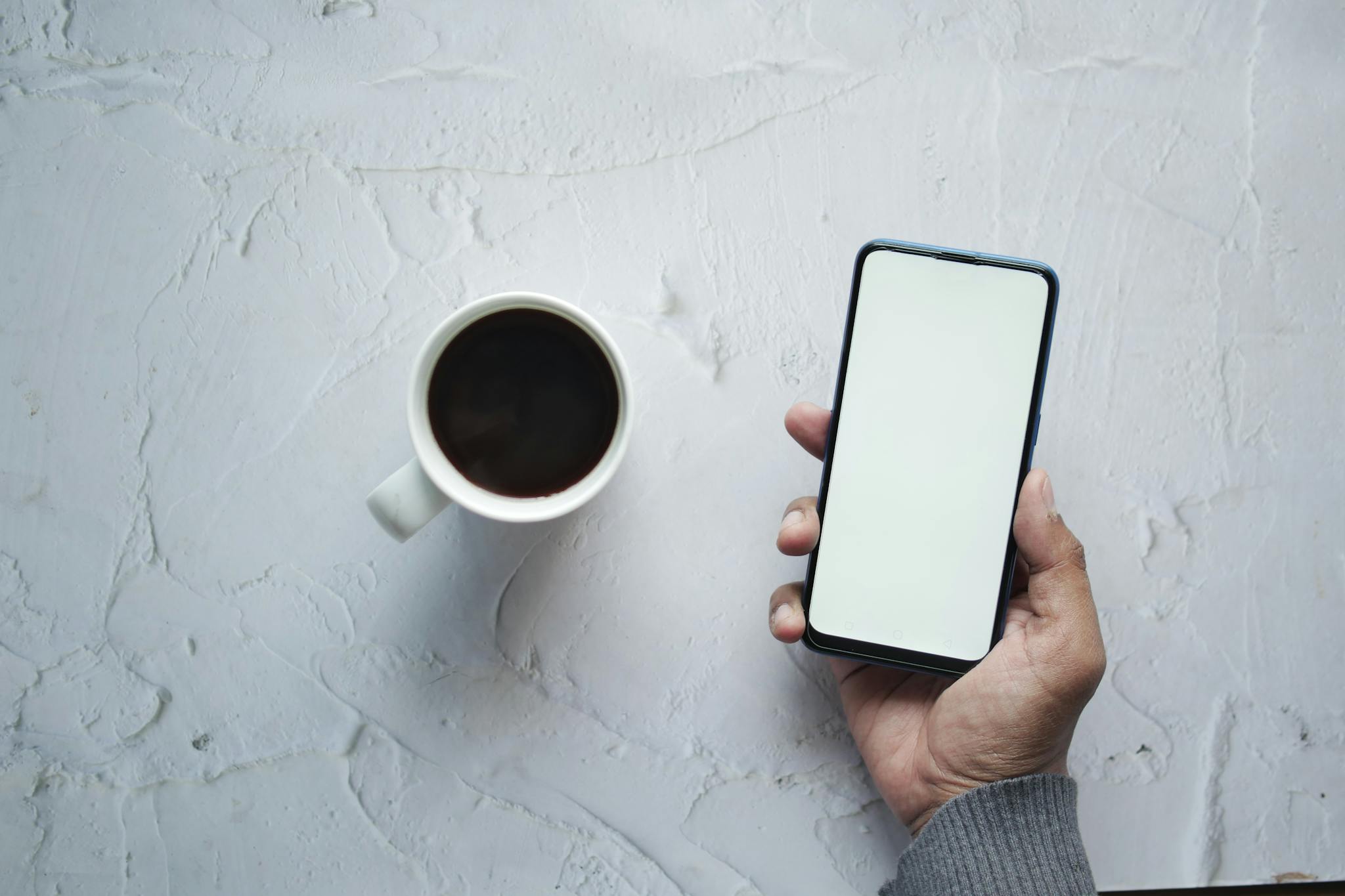 Hand holding smartphone with blank screen next to a cup of coffee on a textured white background.